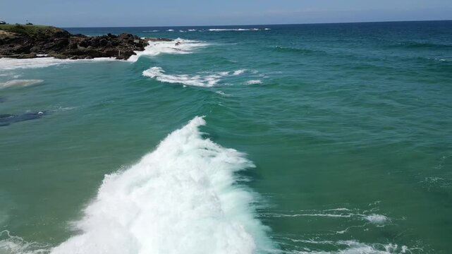Moving fast towards the headland with lots of waves through the blue ocean and waves crashing on the rocks in the background.