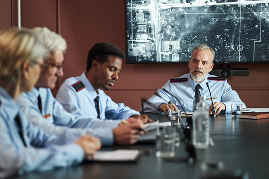 Group of middle aged and senior politicians including Caucasian and Black men and women sitting at conference table, discussing strategy with large map displayed on screen