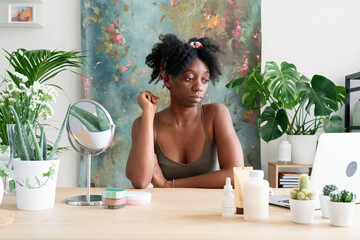 Focused black female sitting at table with various cosmetic products and watching online tutorial on beauty routine treatment via netbook at home