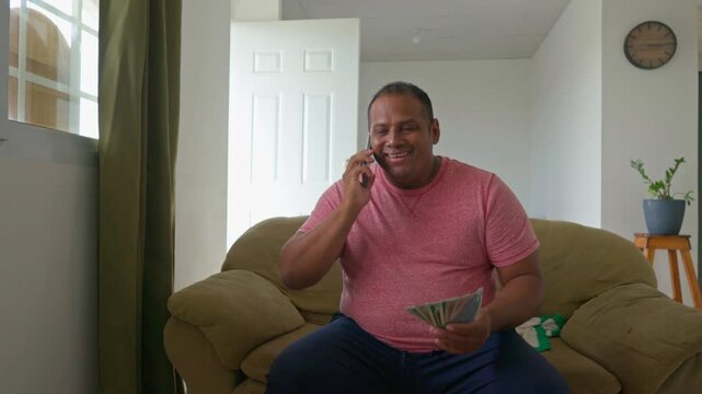 A man sitting on a couch with a cell phone and a stack of money in his hand