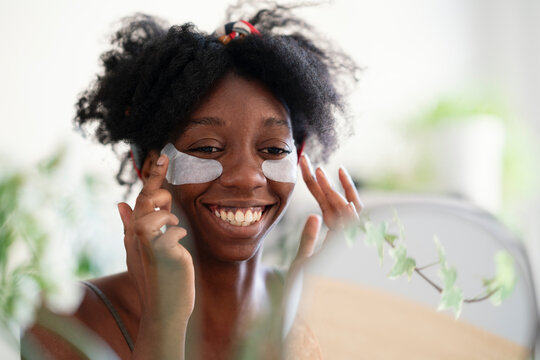 Cheerful black female applying eye patches and doing beauty routine in front of mirror at home
