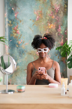Black female in facial sheet mask sitting at table and browsing mobile phone during skincare routine