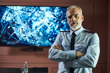 Portrait of middle aged Caucasian man with gray hair and beard standing with arms crossed in front of large digital map display, wearing uniform and looking into camera