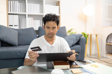 A man is sitting on the floor. He is looking at his credit card and checking his expenses on his...
