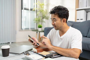 A man is sitting on the floor. He is looking at his credit card and checking his expenses on his...