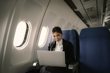 A businesswoman sits in an airplane seat working on a laptop, focused and productive during her...