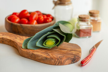 wooden texture cutting board. Nearby lies a knife with a red stylish handle. In the background are tomatoes in a wooden-textured bowl and glass jars of spices.