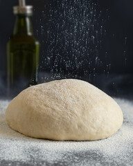 Close-up shot of dough lying on a dark table. There is a bottle of vegetable oil in the back. Flour is pouring.