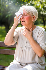 Senior woman feeling unwell from a heatstroke, touching her neck and wiping sweat in a park. depicting symptoms of dehydration or a heart problem in hot weather