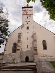 Fototapeta premium Basilica of the Holy Cross in Kežmarok, Slovakia. Historic Roman Catholic church with tall tower, old plaster, and trees around, seen under cloudy sky. 