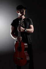 A musician dressed in black holds a guitar while posing calmly in a dimly lit studio. The setting...