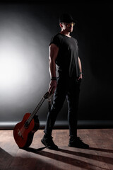 A musician exudes confidence while standing against a dark backdrop, holding a guitar in one hand....