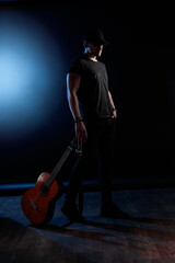 A musician stands confidently in a dimly lit space, wearing a hat and black attire. A guitar leans...