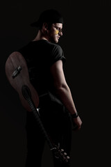 A musician stands confidently with an acoustic guitar resting against his back in a dimly lit...