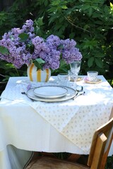 Beautiful lilac flowers, plates, cutlery, glasses and candles on table in garden