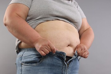 Overweight woman trying to put on tight jeans against grey background, closeup