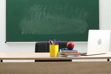 Different school stationery, laptop and red apple on wooden table near blackboard indoors