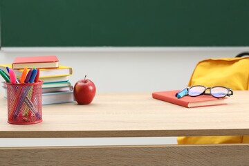 Different school stationery, glasses and red apple on wooden table near blackboard indoors