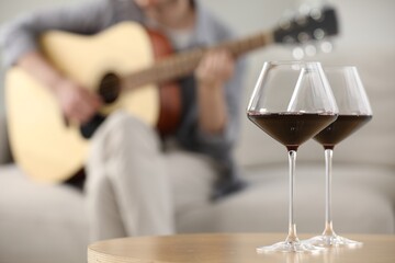 Glasses with red wine on table and man playing guitar indoors, selective focus