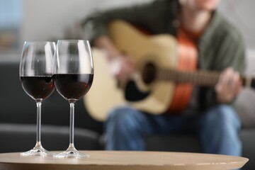 Glasses with red wine on table and man playing guitar indoors, selective focus