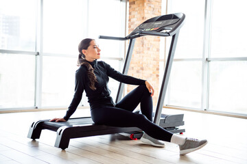 Active woman sitting on a treadmill in a bright gym wearing activewear, embodying a healthy and...