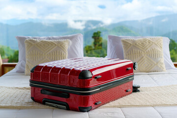 Red suitcase placed on a cozy white bed in a resort room with panoramic forest and mountain view outside glass windows. Perfect start to a relaxing nature getaway.