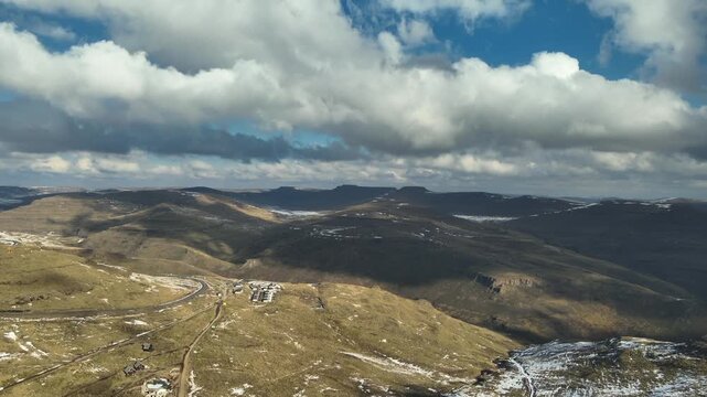 Aerial view of Lesotho&rsquo;s winter mountains shows scattered snow on rugged peaks beneath a bright blue sky, with heavy clouds drifting across the highlands.