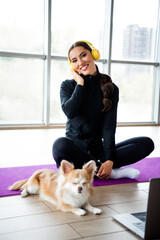Young woman in sportswear enjoying music on headphones while sitting with her dog indoors on a yoga...