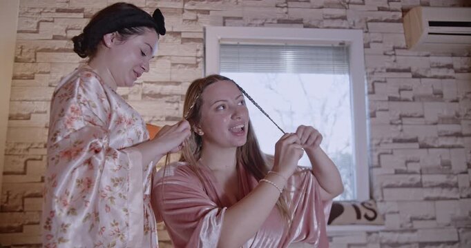 Two young women laugh and have fun as they braid each other's hair in their robes, enjoying a relaxing morning together.