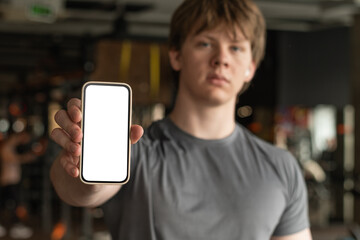 Athlete standing and showing cellphone with white blank screen in blurred gym setting. Fitness App....