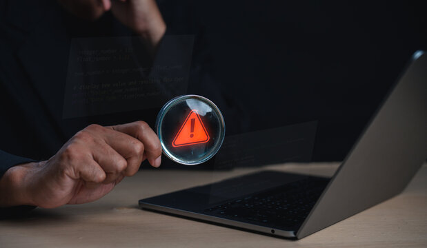 A person holds a magnifying glass highlighting a red warning symbol on a laptop screen in a dark setting.