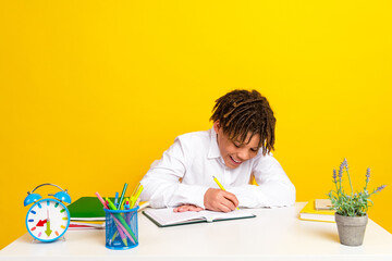 Smiling schoolboy in white shirt writing in notebook at desk with supplies in colorful setting
