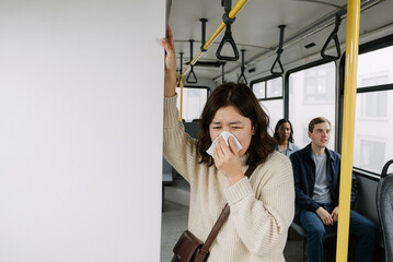 young woman sneezing into tissue on public bus during commute with passengers seated in background wearing casual clothing