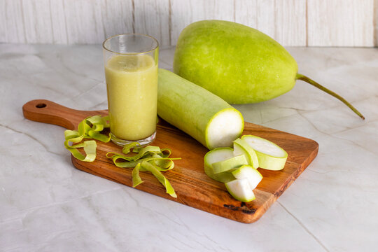 A fresh bottle gourd with slices, peels, and a glass of green juice are arranged on a wooden board over a white marble surface.
