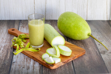 A fresh bottle gourd with slices, peels, and a glass of green juice are arranged on a wooden board...