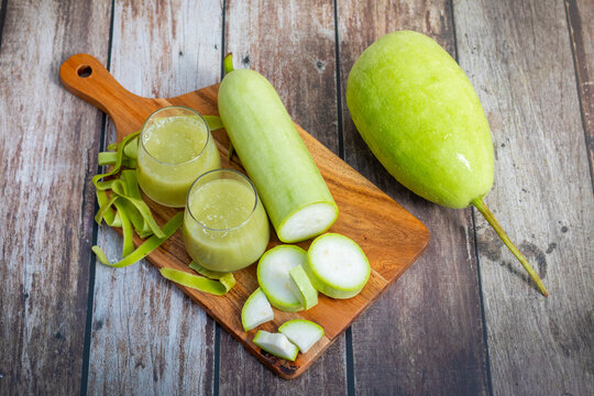 Two glasses of fresh bottle gourd juice, peeled slices, and whole gourd are placed on a wooden board over rustic wooden background, showcasing health and freshness.
