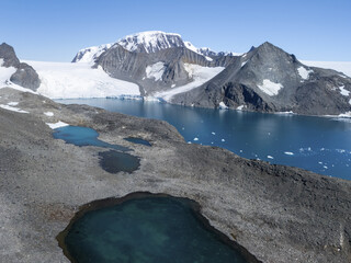 Aerial view of turquoise waters nestled amidst the rugged, rocky landscape, with snow-capped mountains in the distance, Seymour Island, Antarctica.