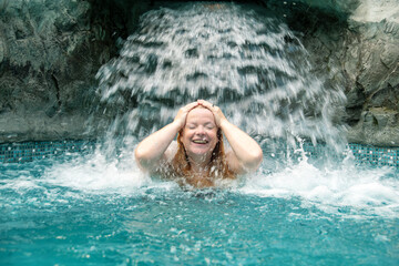 Joyful woman enjoys a refreshing waterfall in a pool, smiling with her hands on her head. Water...