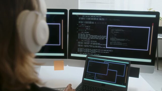 Back shot of unrecognizable blonde woman listening to music while coding via computer, she sitting at desk with two monitors and laptop
