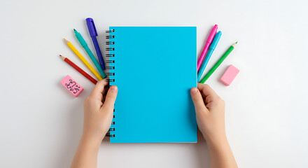 A child&rsquo;s hands holding an empty-cover spiral notebook, with bright school supplies like pencils and pens around, all against a clean white background