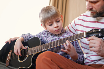 Handsome dad teaches his cute son to play the guitar sitting on sofa in the living room at home....