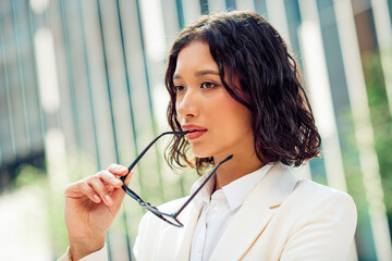Confident young businesswoman in elegant white jacket holding glasses outdoors in urban city street...