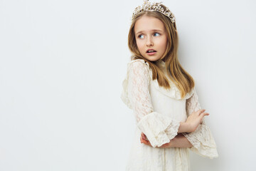 A young girl wearing a white lace dress and tiara, expressing a thoughtful emotion against a soft...