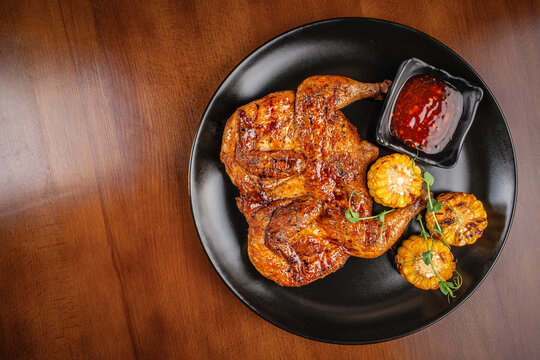 Grilled whole spatchcock chicken served with charred corn rounds and a side of sweet chili sauce on a black plate.