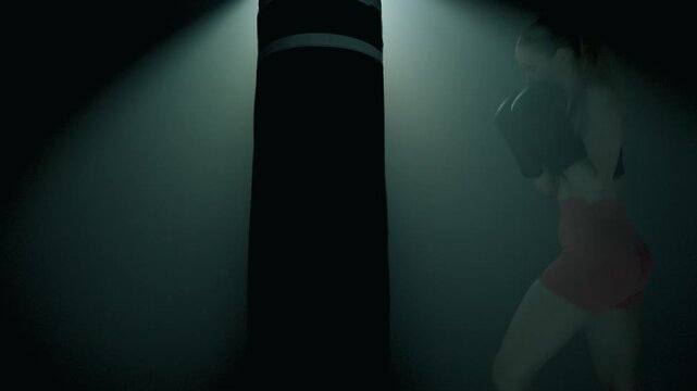 Profil bokserki bijącej mocno w worek w ciemnej sali, światło filmowe padające ku kamerze, sylwetka od dołu, płynne przejście kamery w g&oacute;rę, siła, dynamika, sportowy dramat