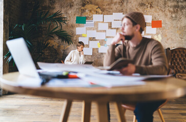 Young freelancer woman analyzes data on laptop near moodboard while man brainstorms with notebook,...
