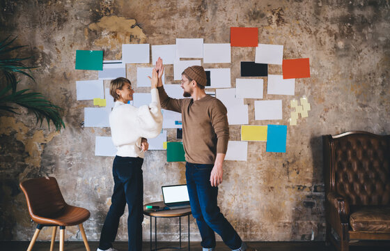 Freelancers share moment of tech success, smiling and connecting palms in front of UX board filled with interface layouts and analytics — visualizing startup completion.