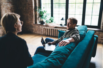 Freelancer sits with laptop on teal couch speaking to woman, showing relaxed modern workflow,...