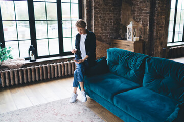 Mature businesswoman uses stylus with smartphone while seated on teal sofa, reflecting mobile...