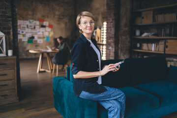 Confident middle-aged freelancer woman using smartphone on couch in creative office, representing...